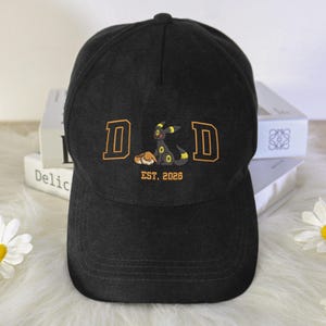 Op de afbeelding: Zwarte baseballcap met geborduurd ontwerp. Het ontwerp bevat de letters "D&D" in oranje, een cartoonfiguur en de tekst "EST. 2026". De cap heeft een gebogen klep en een knoop aan de bovenkant.