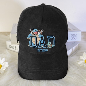 May include: A black baseball cap featuring a graphic print of a blue Blastoise and a small light blue Squirtle positioned around the large blue text DAD, with the smaller blue text EST. 2026 printed directly underneath the main graphic design.