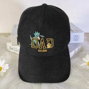 May include: A black baseball cap featuring a graphic print with the word DAD in yellow block letters. A teal creature is positioned above the letters, and a small yellow creature is next to the letter D. The text EST.2026 is printed below.