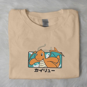 May include: A beige t-shirt with an embroidered orange dragon design, teal wings, and Japanese text "カイリュー". The dragon is set within a rectangular frame.