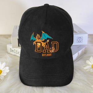May include: Black baseball cap with an embroidered design featuring a cartoon dragon, the word "DAD" in orange block letters, and the text "EST.2026". The cap has a curved brim and a button on top.