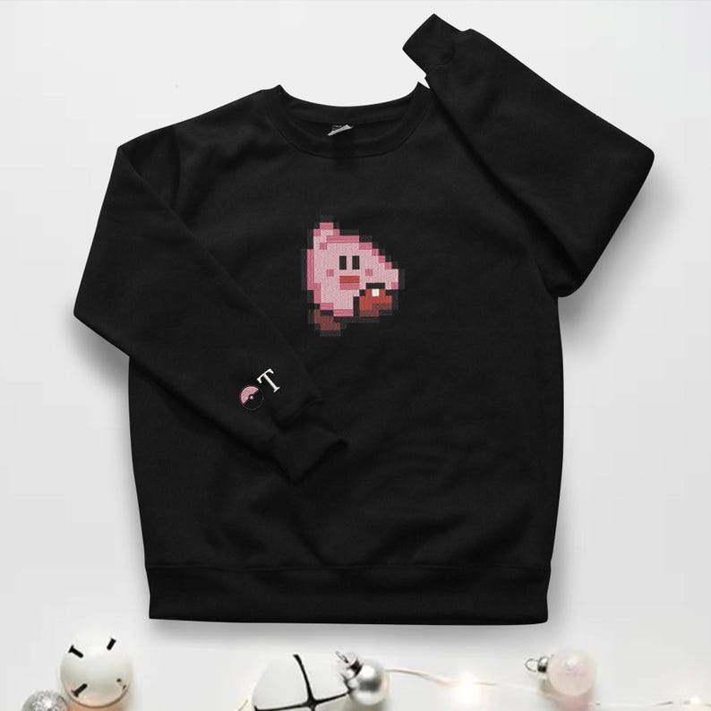May include: Black crew neck sweatshirt with a pixelated pink character design. The left sleeve has a small graphic with the letters "T". The sweatshirt is a solid black colour.