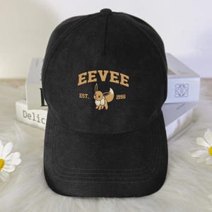 以下が含まれることがあります： 黒い野球帽。 金色の文字で「EEVEE」と刺繍されたイーブイのキャラクターと「EST. 1996」の文字が特徴です。 キャップはカーブしたつばと丸いクラウンが付いています。