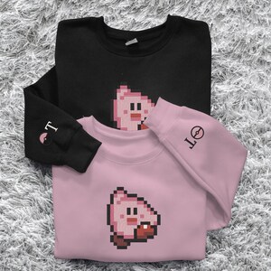 May include: Two sweatshirts, one black and one pink, are stacked on a textured surface. Each sweatshirt features a pixelated cartoon character on the front. The black sweatshirt has "T" and a symbol on the sleeve, while the pink one has "OT" on the sleeve.