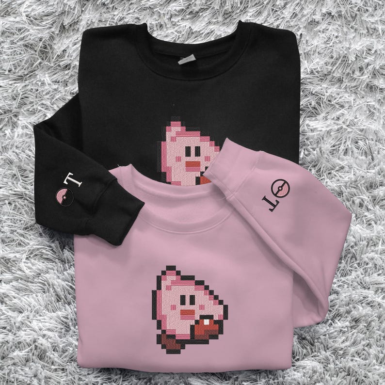 May include: Two sweatshirts, one black and one pink, are stacked on a textured surface. Each sweatshirt features a pixelated cartoon character on the front. The black sweatshirt has "T" and a symbol on the sleeve, while the pink one has "OT" on the sleeve.