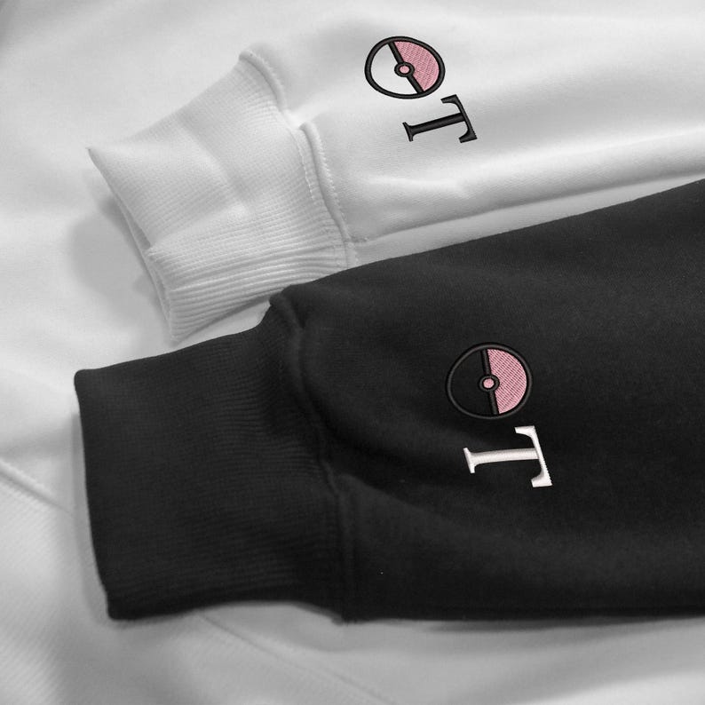May include: Close-up of two sweatshirts, one white and one black. Both have a circular embroidered design with a pink centre and the letter "T" in white. The cuffs are ribbed.