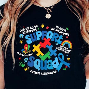 Op de afbeelding: Zwart T-shirt met een kleurrijk grafisch ontwerp met de tekst "Support Squad" en inclusief puzzelstukjes, een hart, een vlinder, een regenboog en de tekst "It's ok to be different", "Neurodiversity", "Everyone communicates differently", "In April we wear blue" en "Autism Awareness".
