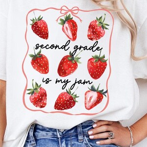 Può includere: T-shirt bianca con un design a fragole e la scritta "second grade is my jam". Il design presenta fragole rosse e un bordo rosa. Un capo casual per tutti i giorni.