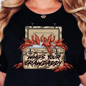 May include: Black t-shirt featuring a camouflage cooler graphic filled with red crawfish and ice. The text "WHO'S YOUR CRAWDADDY" is printed below the cooler. The shirt is a casual wear item.
