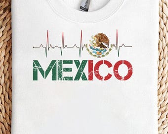 May include: White sweatshirt featuring the word "MEXICO" in green and red, with a heartbeat line design above it. The Mexican coat of arms is on the right side of the heartbeat line. The sweatshirt is on a woven, circular surface.