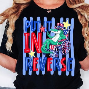 以下が含まれることがあります： 黒いTシャツで、愛国的なシルクハットとサングラスを着用し、車椅子に座っている緑色のワニのグラフィックが描かれています。テキストは「PUT IT IN REVERSE」と赤と青の文字で書かれ、青い縦縞と黄色い星で囲まれています。