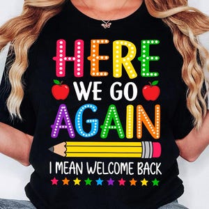 May include: A black t-shirt featuring the colorful text "HERE WE GO AGAIN I MEAN WELCOME BACK" in a dotted font. The design includes two red apples, a yellow pencil with a pink eraser, and a row of small multi-colored stars at the bottom.
