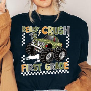 Pode incluir: Camiseta preta com um gráfico de um monster truck de camuflagem verde. O texto "READY TO CRUSH FIRST GRADE" é exibido em laranja, verde e branco. O design inclui um padrão quadriculado.
