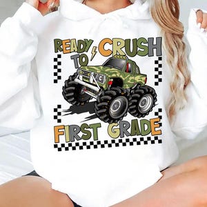 Peut inclure: Sweat à capuche blanc avec un motif de camion monstre camouflage et le texte "READY TO CRUSH FIRST GRADE". Le design comprend un éclair et des accents de drapeau à damier. Le texte est en orange, vert et noir.