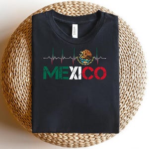 May include: Black t-shirt with the word "MEXICO" in green, white, and red, and a heartbeat line design. The Mexican coat of arms is also featured. The t-shirt is folded on a woven surface.