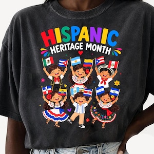May include: A charcoal gray t-shirt featuring a colorful graphic with the text "HISPANIC HERITAGE MONTH" in bold letters. The design shows six cartoon children in traditional clothing holding various national flags, surrounded by small flowers.