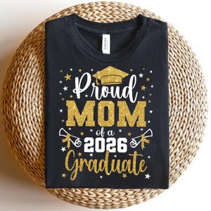 May include: Black t-shirt with gold glitter text that reads "Proud MOM of a 2026 Graduate." The design includes a graduation cap, stars, and scrolls. The shirt is displayed on a woven, circular surface.