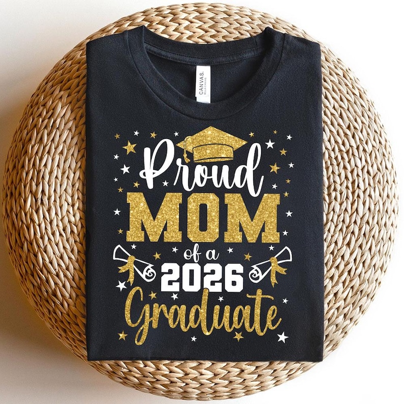 May include: Black t-shirt with gold glitter text that reads "Proud MOM of a 2026 Graduate." The design includes a graduation cap, stars, and scrolls. The shirt is displayed on a woven, circular surface.