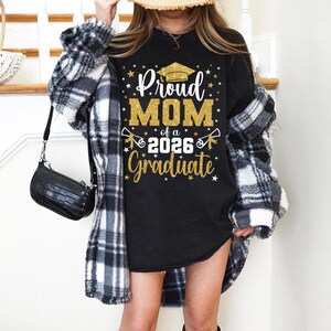 May include: Black t-shirt with gold glitter text that reads "Proud MOM of a 2026 Graduate" with a graduation cap graphic. The shirt is worn with a black and white plaid jacket and a black purse.