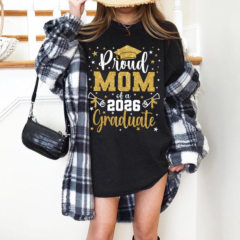 May include: Black t-shirt with gold glitter text that reads "Proud MOM of a 2026 Graduate" with a graduation cap graphic. The shirt is worn with a black and white plaid jacket and a black purse.