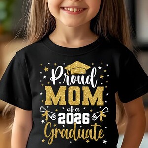May include: Black t-shirt with gold glitter text that reads "Proud MOM of a 2026 Graduate." The design includes a graduation cap, stars, and diploma illustrations. A child is wearing the shirt.