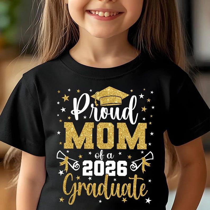 May include: Black t-shirt with gold glitter text that reads "Proud MOM of a 2026 Graduate." The design includes a graduation cap, stars, and diploma illustrations. A child is wearing the shirt.