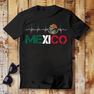 May include: Black t-shirt with the word "MEXICO" in green, white, and red, and a heartbeat graphic. The Mexican coat of arms is above the word. Sunglasses are on a wooden surface.
