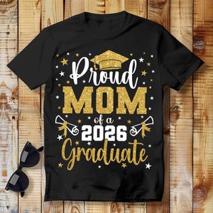 May include: Black t-shirt with the text "Proud MOM of a 2026 Graduate" in white and gold glitter lettering. The design includes a graduation cap, stars, and diploma illustrations. Sunglasses rest on the wooden surface.