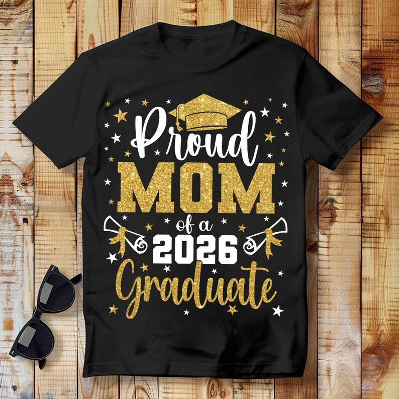 May include: Black t-shirt with the text "Proud MOM of a 2026 Graduate" in white and gold glitter lettering. The design includes a graduation cap, stars, and diploma illustrations. Sunglasses rest on the wooden surface.