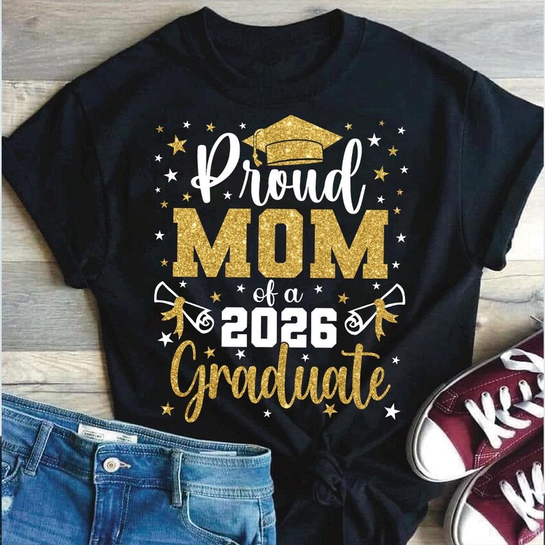 May include: Black t-shirt with a gold glitter design that reads "Proud MOM of a 2026 Graduate." The design includes a graduation cap, stars, and diploma scrolls. Blue jeans and maroon sneakers are visible in the image.