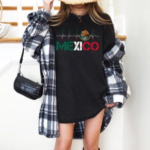 May include: Black long-sleeve shirt with the word "MEXICO" in green, white, and red, with a heartbeat line design. The shirt features the Mexican coat of arms. The person is wearing a black and white plaid shirt.