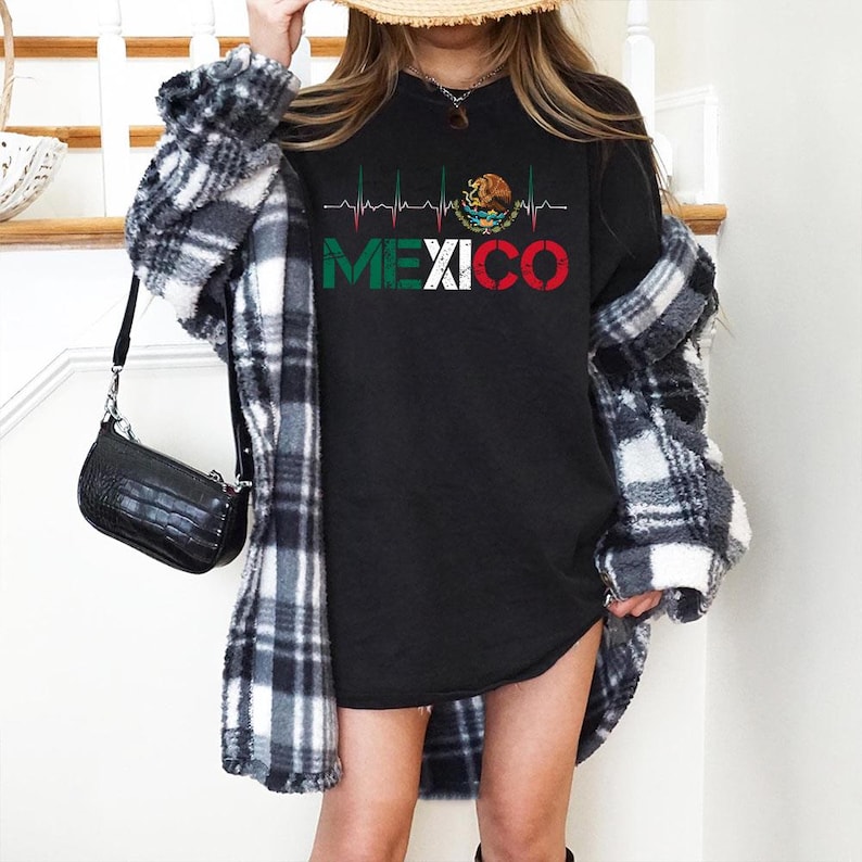 May include: Black long-sleeve shirt with the word "MEXICO" in green, white, and red, with a heartbeat line design. The shirt features the Mexican coat of arms. The person is wearing a black and white plaid shirt.