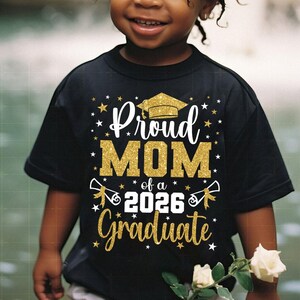 May include: Black t-shirt with gold glitter text that reads "Proud MOM of a 2026 Graduate." The design includes a graduation cap and stars. The shirt is worn by a child.