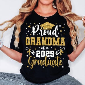 May include: Black t-shirt with gold glitter text reading "Proud Grandma of a 2025 Graduate." The design features a graduation cap, stars, and scrolls. The shirt has a crew neck.