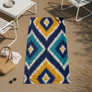 May include: A rectangular beach towel with a geometric ikat pattern in navy blue, teal, and yellow. The towel is lying on a light brown surface, with white chairs and a straw bag in the background.