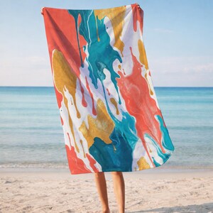May include: A beach towel with a colorful abstract design. The towel features a white background with splashes of gold, teal, red, and orange paint. The towel is being held up on a sandy beach with the ocean in the background.