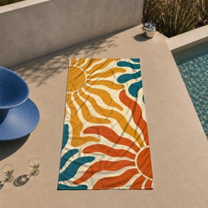 May include: A rectangular beach towel featuring a retro sunburst design. The towel has a cream-colored background with large sun shapes in orange, yellow, and teal. The towel is lying on a concrete surface near a pool.