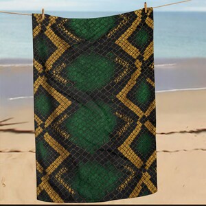 May include: A green, black, and gold patterned beach towel hanging on a clothesline. The towel features a snake skin design with a repeating pattern of green diamond shapes, black scales, and gold diagonal lines. The background shows a beach and ocean.