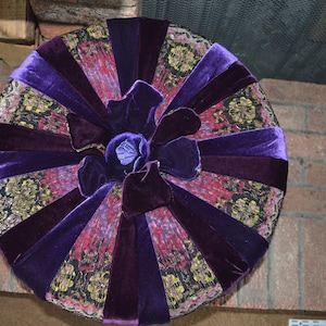 May include: A round purple and gold cushion with a large purple flower in the center. The cushion is made of velvet and has a patterned fabric.