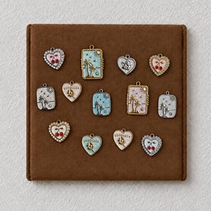 Könnte beinhalten: Eine Sammlung von dreizehn Metallanhängern mit verschiedenen Motiven wie Herzen, Rechtecken, Palmen, Kirschen und dem Schriftzug GOODLUCK. Die Anhänger sind in Gold- und Silbertönen gehalten und mit farbigen Emaille-Akzenten versehen.