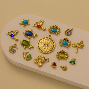 Könnte beinhalten: Eine Sammlung goldfarbener Charms in verschiedenen Formen, darunter Schildkröten, Vögel, Krabben und eine Libelle. Einige Charms weisen türkisfarbene, blaue, rote und grüne Akzente auf. Die Charms sind auf einer hellen Oberfläche angeordnet.
