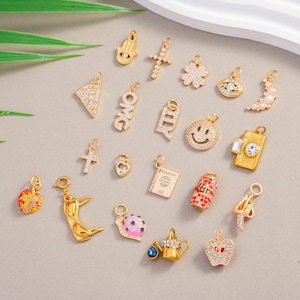 May include: A selection of gold-coloured charms with diverse designs, such as a cross, clover, smiley face, camera, and apple. Some charms are adorned with crystals and pearls, and others feature the words "OMG" and "ILY".