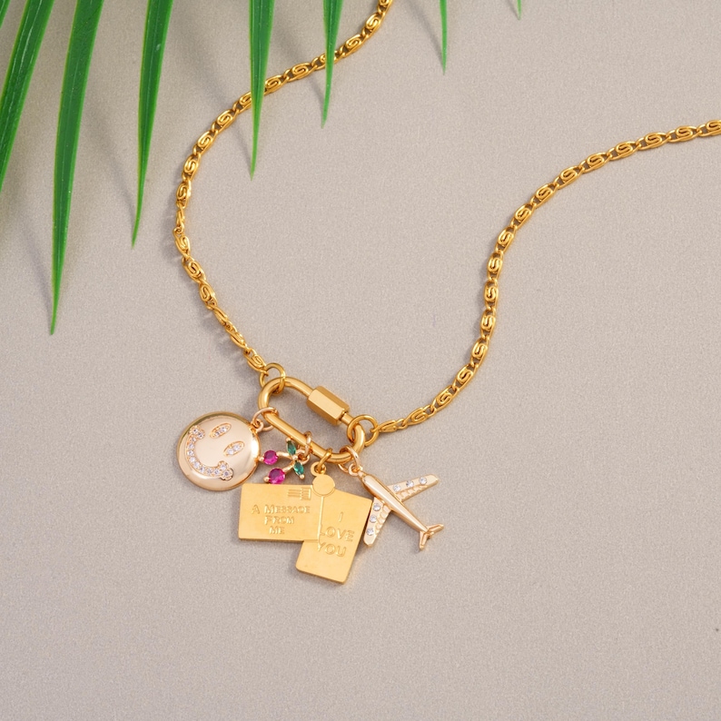 May include: Gold necklace featuring a series of charms. The charms include a smiley face, a small rectangle with the text "A memory next to me", a rectangle with the text "Love you", and a small airplane. The necklace is set against a grey backdrop.