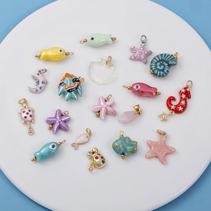 May include: A collection of colorful, sea-themed charms in various shapes, including fish, starfish, and seahorses. The charms are in shades of yellow, pink, blue, and green, with gold-colored accents. The charms are arranged on a white surface.