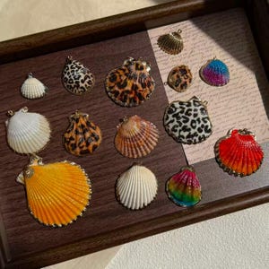 Puede incluir: Una colección de dijes de conchas marinas con varios patrones y colores, incluyendo estampado de leopardo, blanco sólido, amarillo brillante, degradado de rojo a naranja y tonos arcoíris. Cada concha tiene una pequeña argolla dorada.