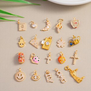 May include: A selection of gold-coloured charms featuring diverse designs such as a passport, heart, crescent moon, apple, Hamsa hand, pizza slice, camera, four-leaf clover, teapot, soda can, snail, cross, smiley face, evil eye, and the letters "OMG" and "ILY".