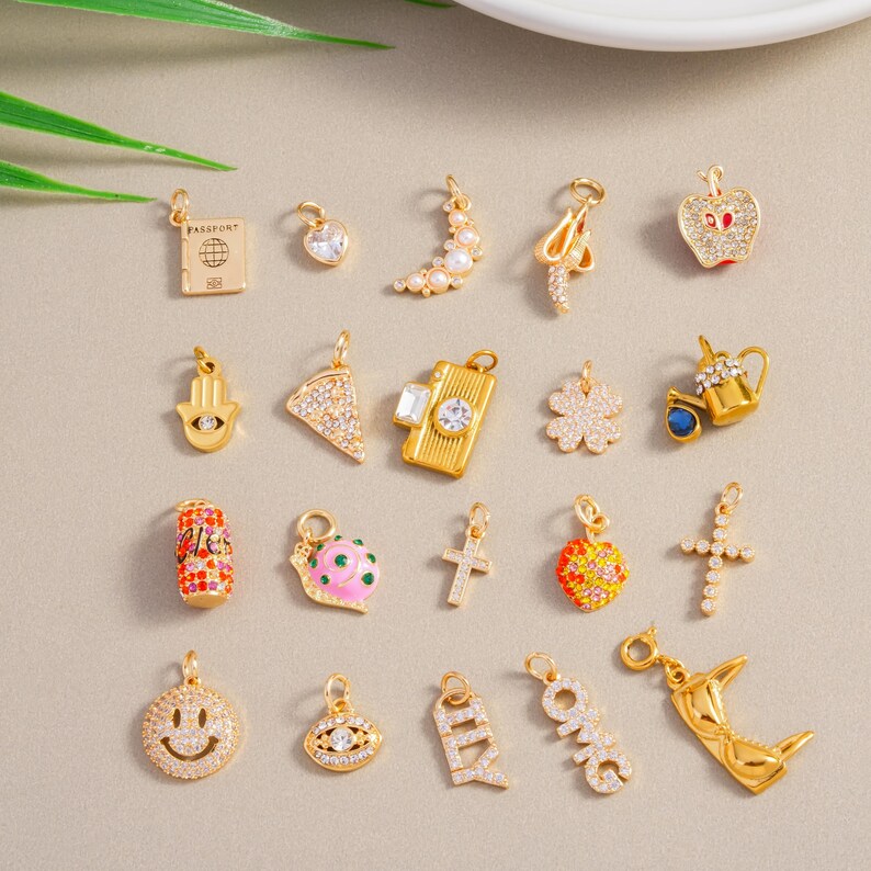 May include: A selection of gold-coloured charms featuring diverse designs such as a passport, heart, crescent moon, apple, Hamsa hand, pizza slice, camera, four-leaf clover, teapot, soda can, snail, cross, smiley face, evil eye, and the letters "OMG" and "ILY".