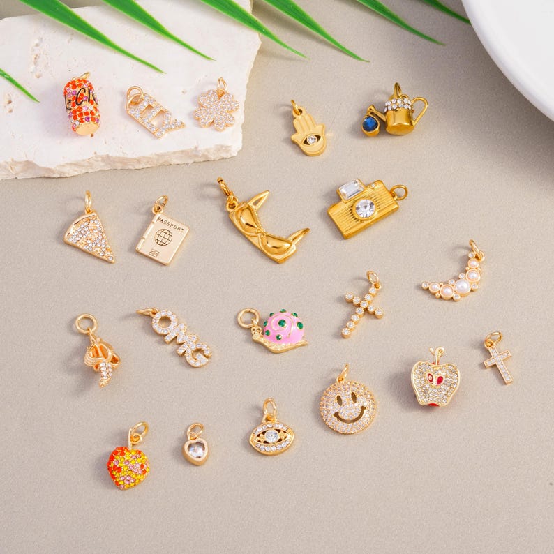 May include: A selection of gold-coloured charms featuring diverse designs such as a camera, a pizza slice, a passport, a smiley face, a cross, and a crescent moon. Some charms are adorned with clear crystals and colourful enamel.