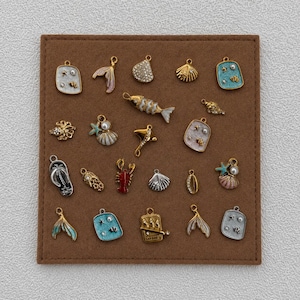 May include: A collection of twenty-one metal charms featuring nautical and ocean-themed designs, including seashells, a fish, a lobster, a flip-flop, and mermaid tails, finished in gold and silver tones with enamel accents and small embedded pearls.
