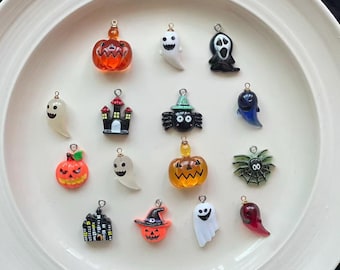 May include: A collection of Halloween-themed charms in various shapes, including pumpkins, ghosts, spiders, and a haunted house. The charms are in orange, black, white, and green, with gold-colored loops for attaching to jewelry or crafts.